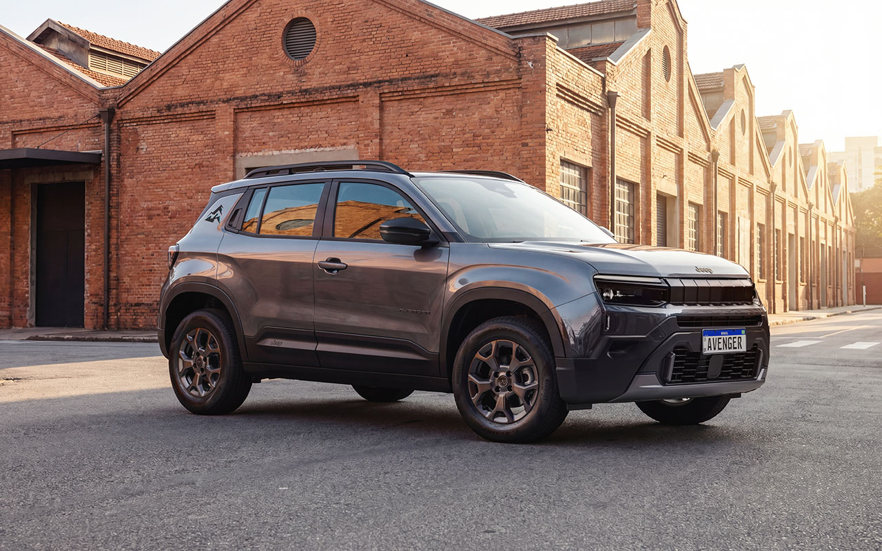 Jeep Avenger Altitude cinza visto de três quartos dianteiros diante de galpões de tijolos