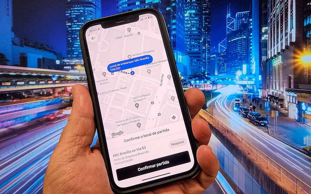Mão segura celular com aplicativo de transporte diante de imagem de vias e edifícios