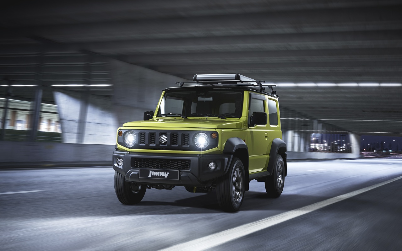 Suzuki Jimny verde a combustão em movimento, visto de frente em três quartos, com faróis acesos